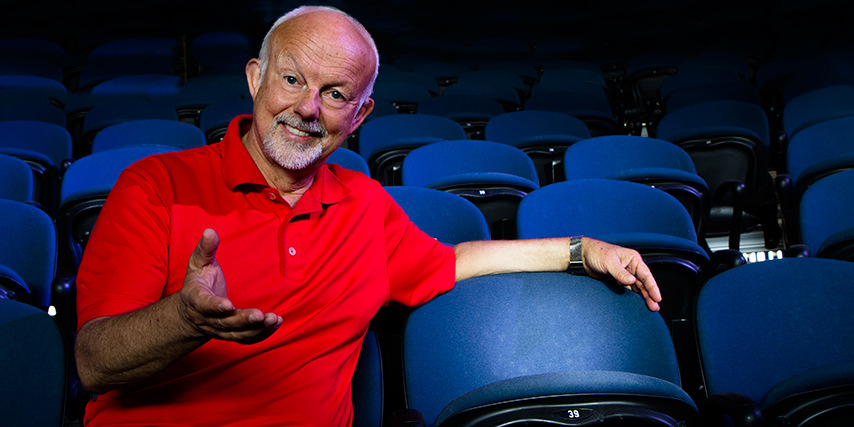 Christopher Gaze, Artistic Director, sitting on a theatre seat with a smile and stretched out hand