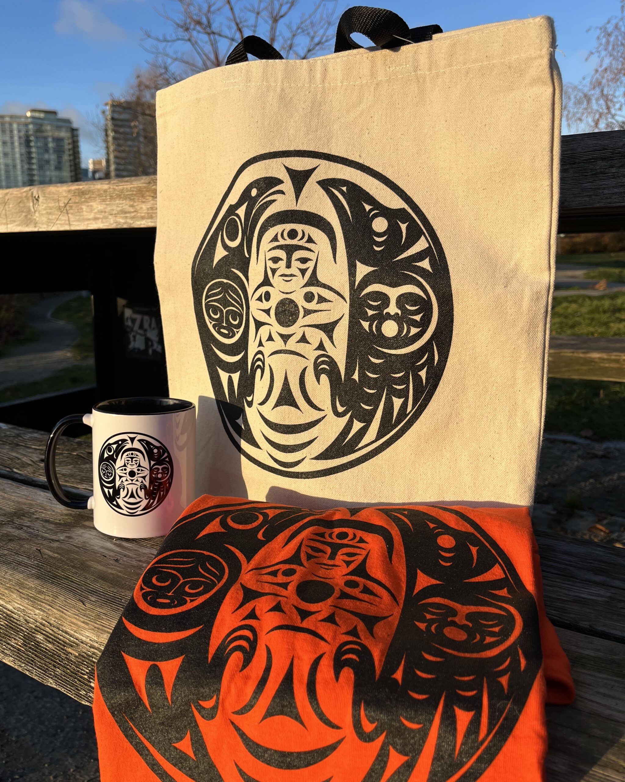 a canvas tote bag, a black and white mug, and an orange shirt arranged on a picnic bench. Each item features the To Learn design