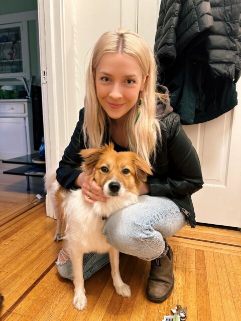 Emma, wearing a black jacket and acid wash jeans, kneels next to her dog Cricket, a medium-sized brown and white dog.