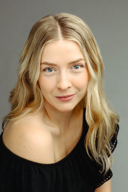 Emma Houghton smiles at the camera wearing a black off-the-shoulder top.