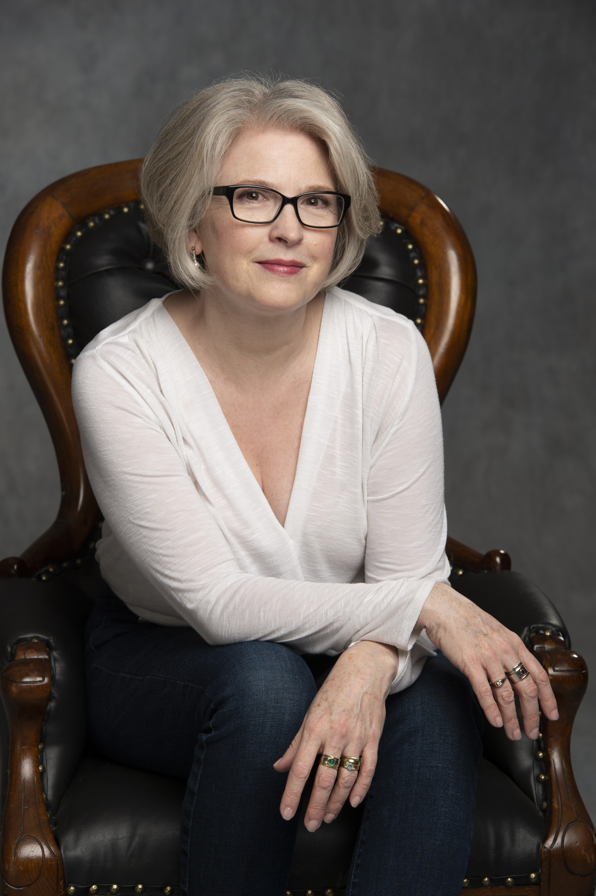 Johnna Wright sits on a leather high-back chair. She wears black glasses, a white blouse, and dark jeans.