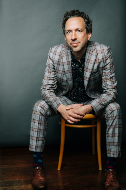 Mark Chavez sits on a stool, leaning forward and wearing a grey checkered suit.