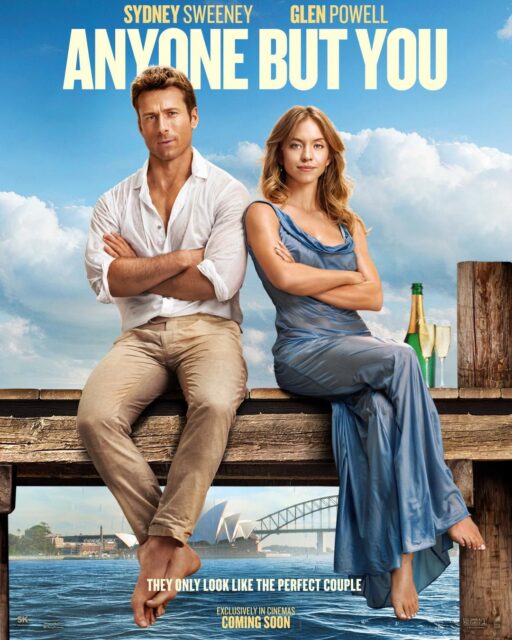 The theatrical release poster for "Anyone But You" (2023). Glen Powell left, and Sydney Sweeney, right, sit shoulder-to-shoulder on a dock with their feet dangling. Powell is wearing khaki pants and a white button-down shirt. His arms crossed and his legs are crossed at the ankles, and he has a half-smirk on his face, looking at the camera. Sweeney has her blonde hair down, past shoulder length, and wears a sky blue, silk, floor-length dress. Her arms and legs are crossed, and she is smirking. There's a bottle and two flutes of champagne beside her. In the background, there is blue sky and fluffy, white clouds, the ocean and the Sydney Opera House and Harbour Bridge.